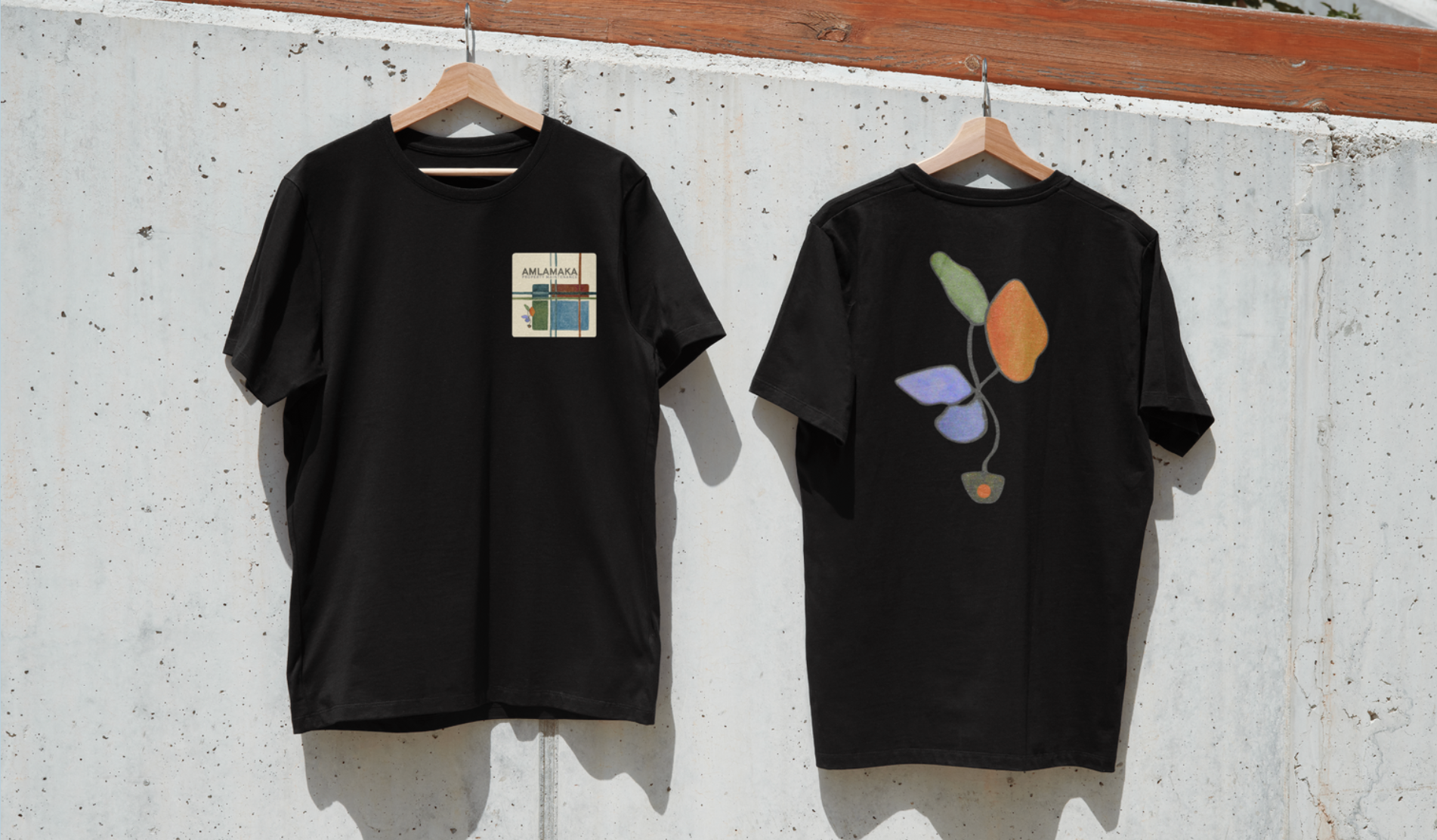 Black AMLAMAKA t-shirts, front chest logo and back plant illustration, hanging on a concrete wall