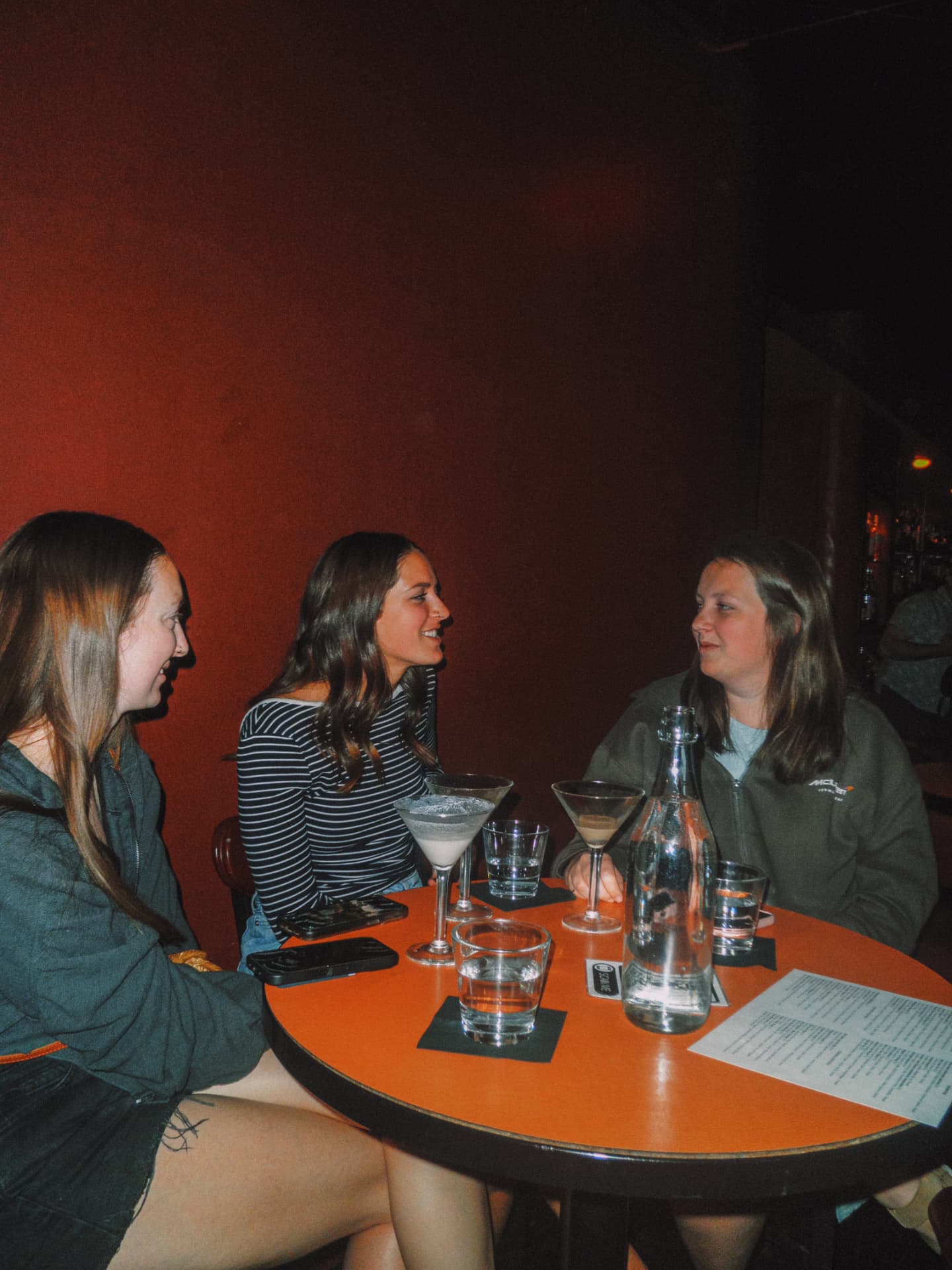 Three friends talking over martinis at an orange table against the red wall