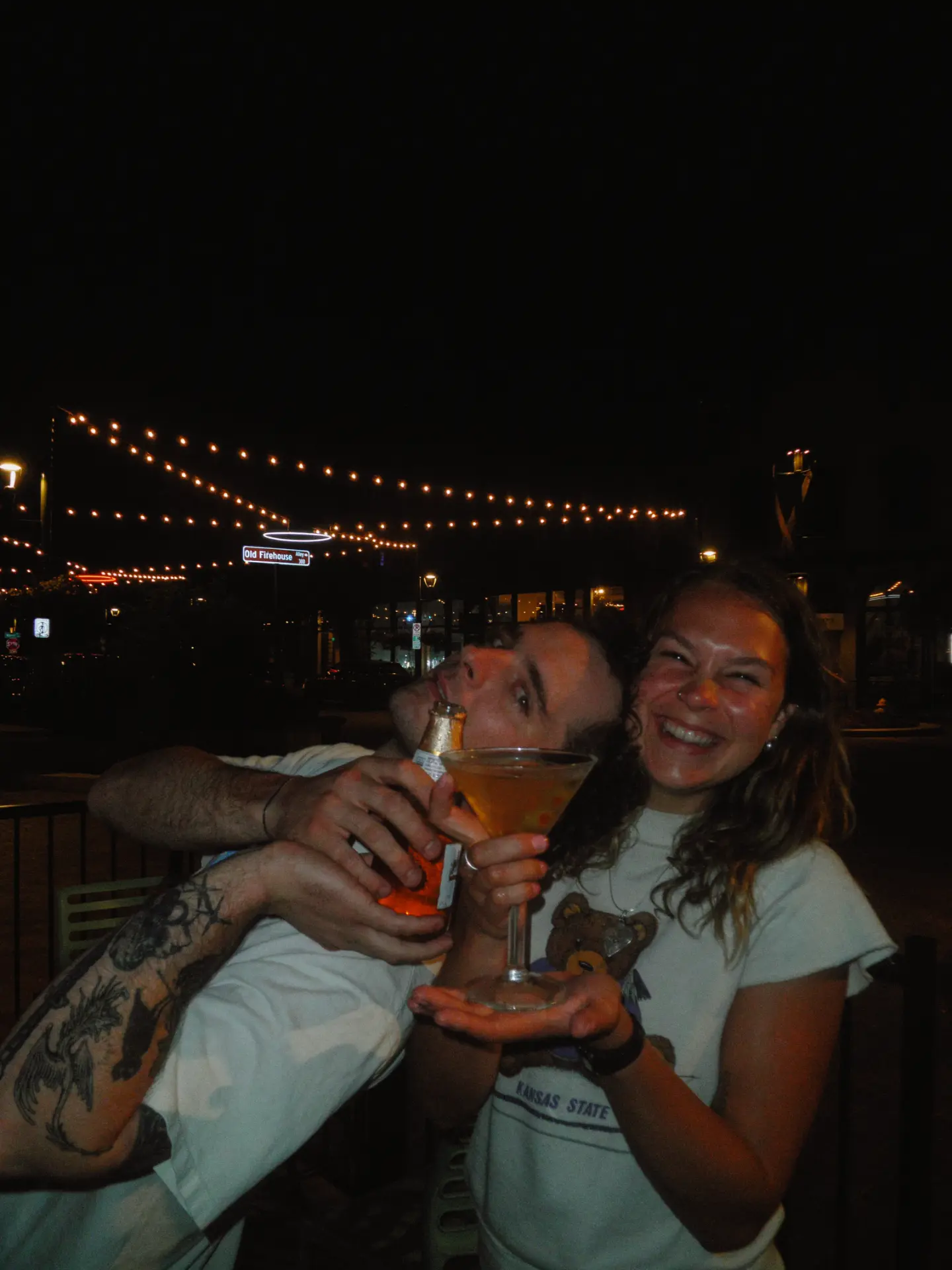 Two guests laughing on the patio at night, sharing a martini under string lights