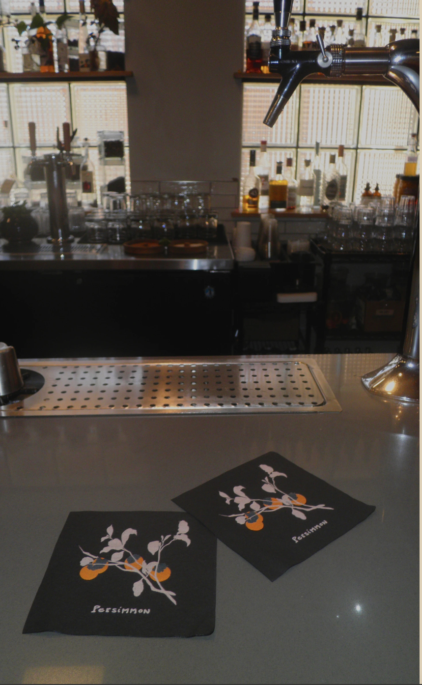 Black cocktail napkins with persimmon branch illustration on the bar top