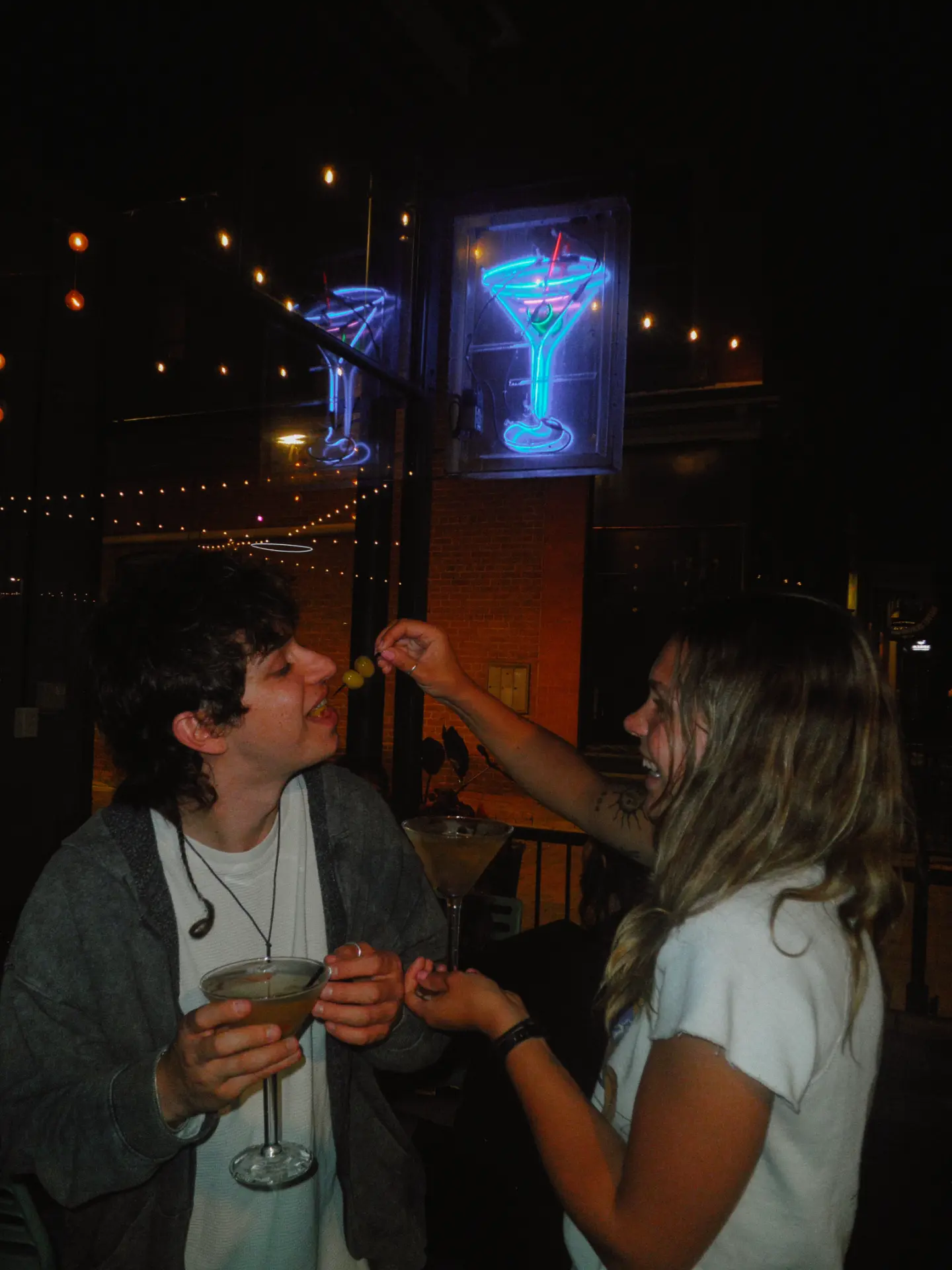 Guest feeding an olive skewer to a friend under the neon martini sign